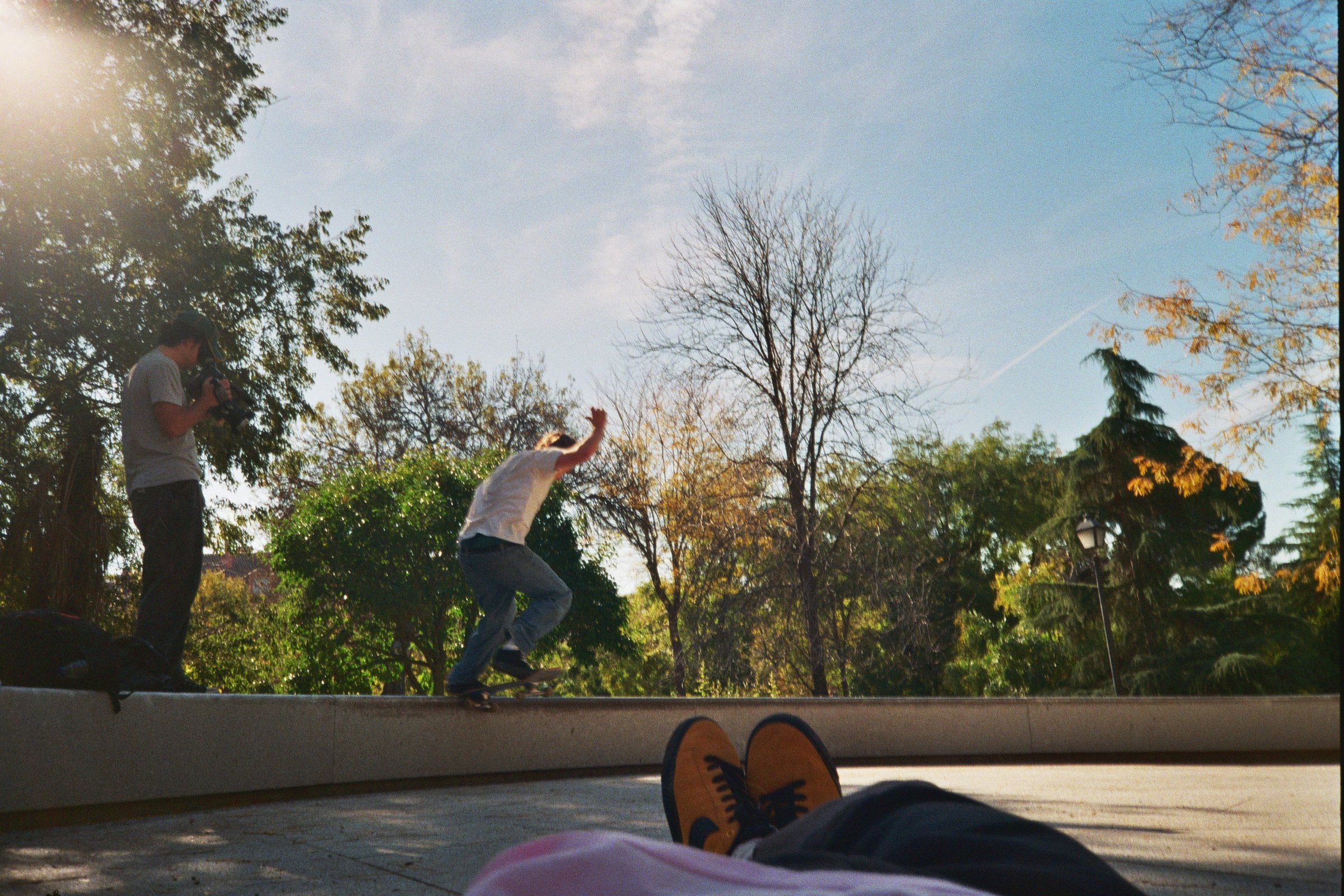 Joven haciendo skateboarding en un parque con árboles y un cielo despejado. Se ve a una persona grabando y otra en medio de la acción, con zapatos deportivos visibles en primer plano.