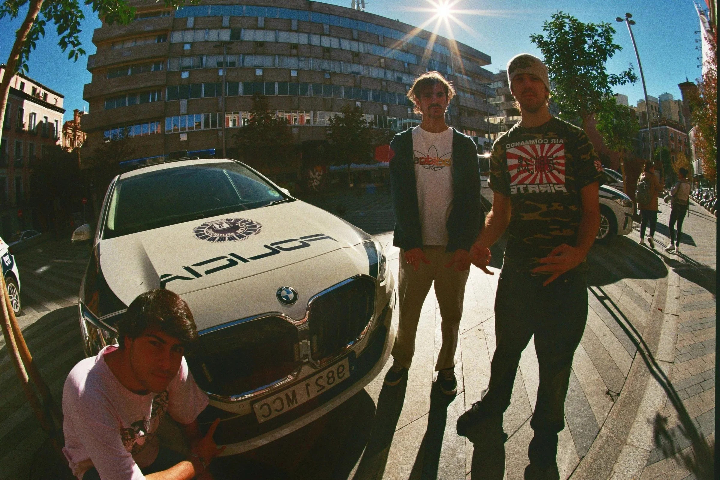Cuatro jóvenes junto a un automóvil de policía en una calle urbana, con edificios y árboles al fondo, y el sol brillando en el cielo.