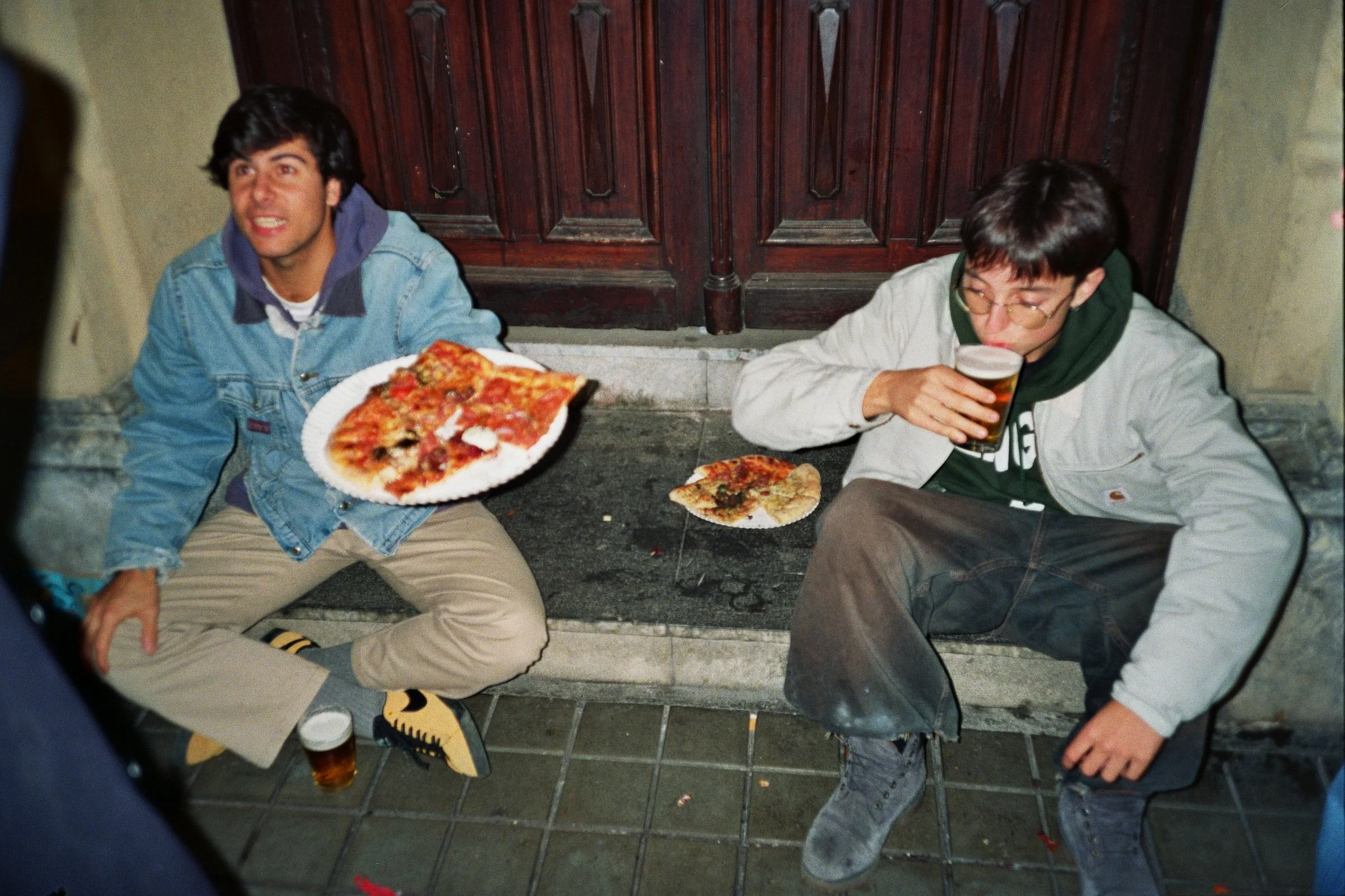 Dos jóvenes sentados en la acera frente a una puerta de madera, uno con una pizza en mano y el otro bebiendo cerveza.