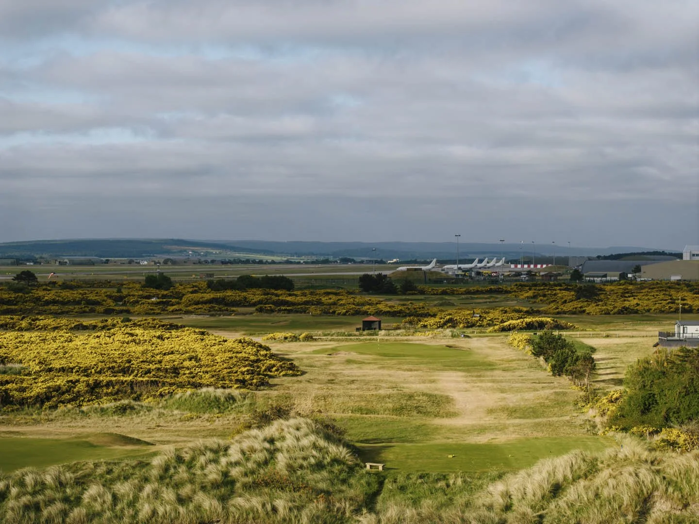A golf course with yellowish-green grass and sand traps in the foreground, rolling hills and trees in the middle ground, and an airport with parked airplanes and airport structures in the background under cloudy skies.