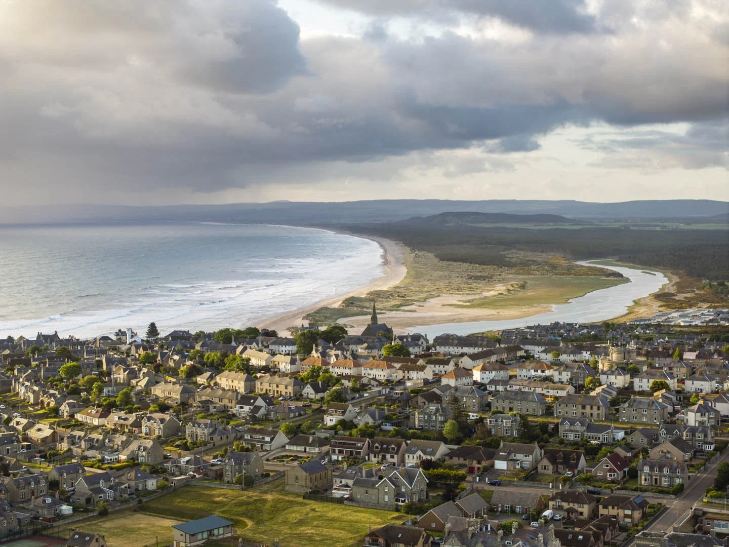 A coastal town with a beach, houses and a church, with a river flowing towards the sea and hills in the background.