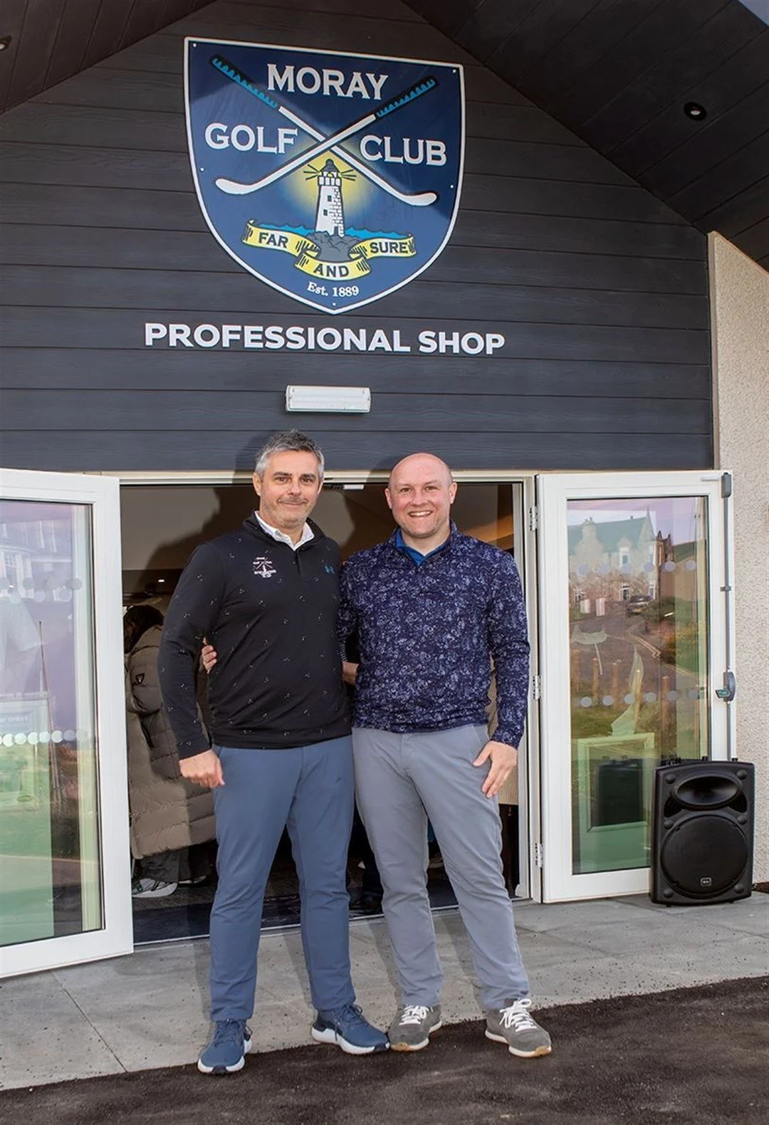 Two men standing in front of the Moray Golf Club professional shop, smiling at the camera. The club's sign is visible above them.