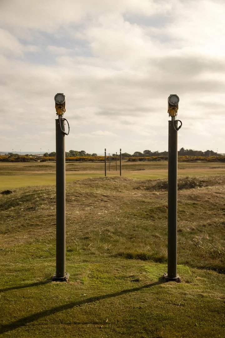 Two large floodlights on tall poles on a grassy hilltop overlooking a golf course or open field, with overcast sky and some distant trees and poles in the background.