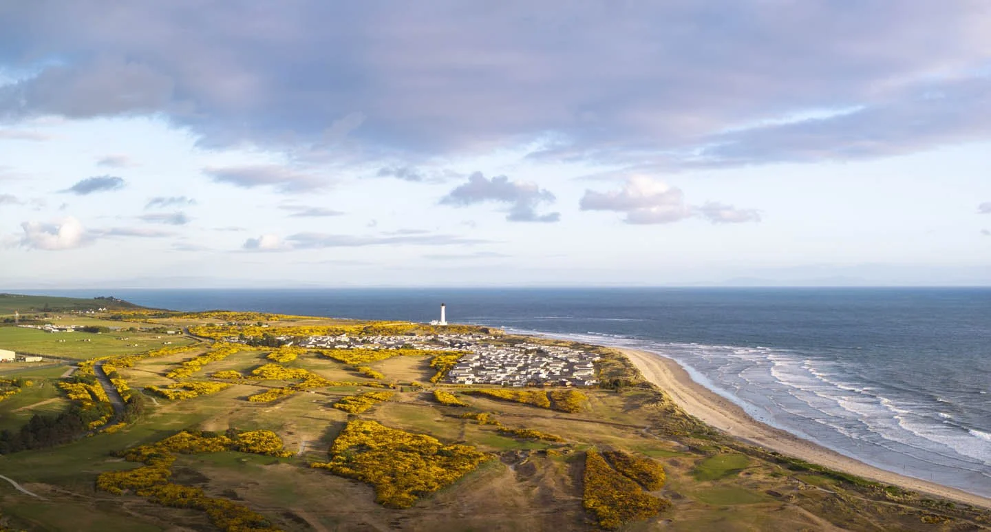 A scenic coastal landscape with a sandy beach along the ocean, a lighthouse in the distance, and rolling fields with yellow flowering bushes in the foreground under a partly cloudy sky.