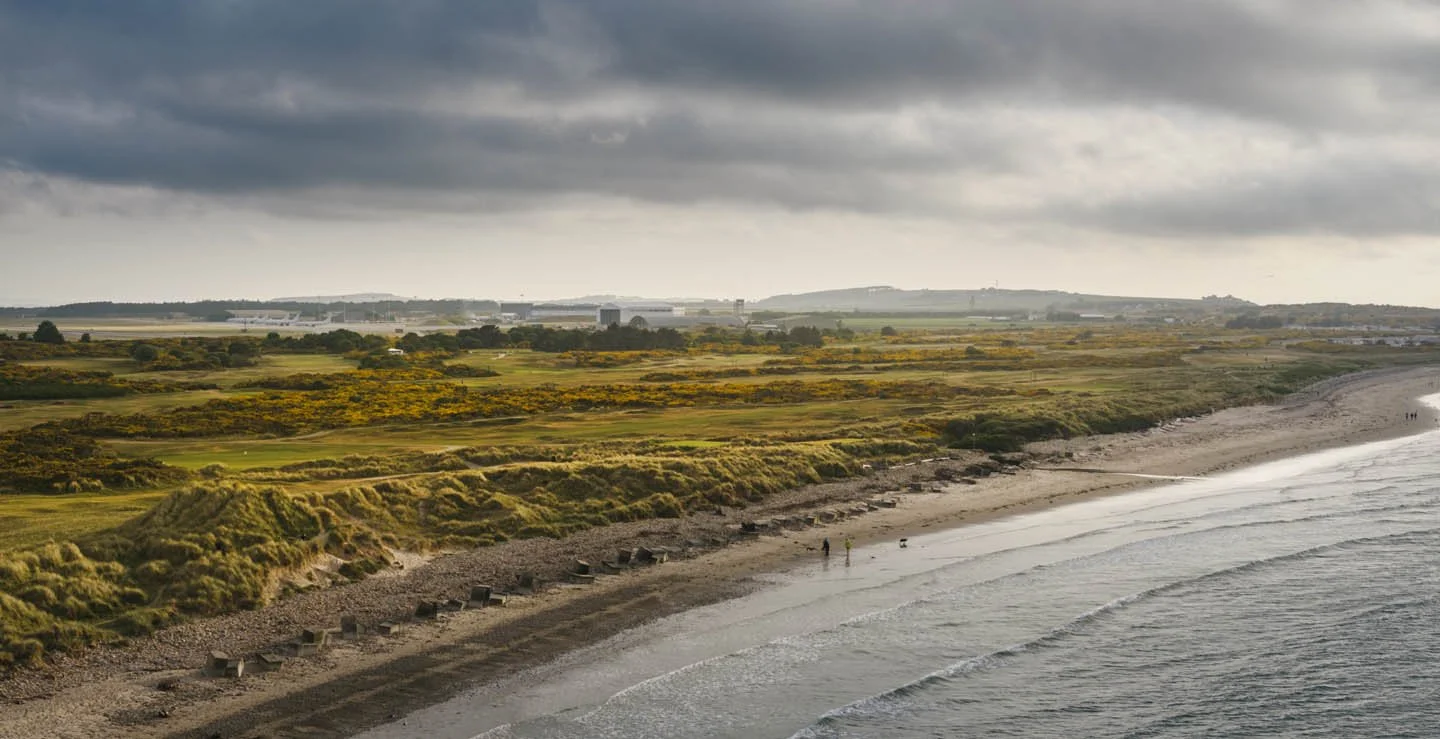 A coastal scene with a sandy beach, grassy dunes, and a cloudy sky. There are a few people walking along the shoreline, and some buildings are visible in the distance.