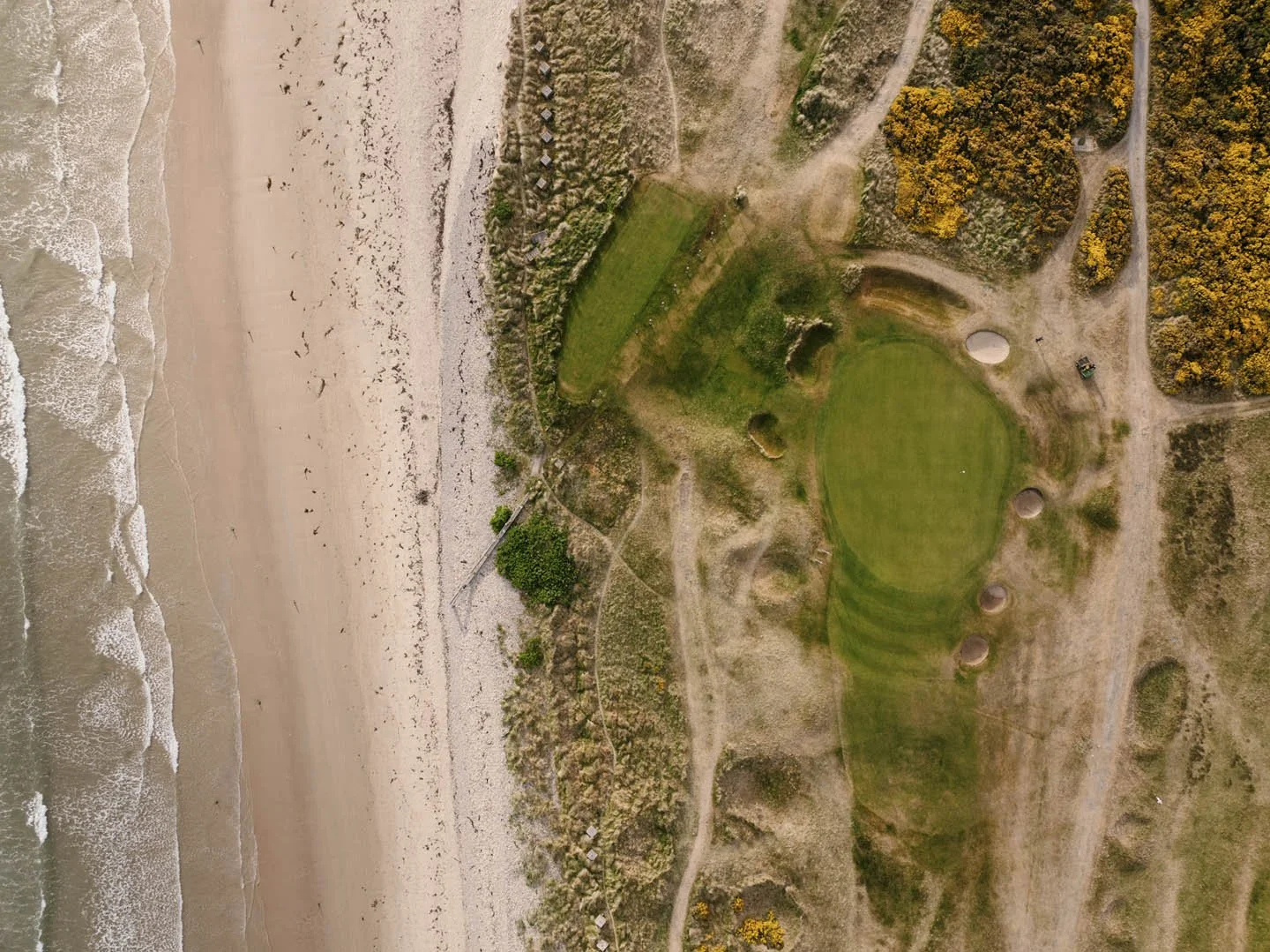 An aerial view of a beach with waves on the left and a grassy golf course with bunkers on the right, surrounded by dirt paths and trees.