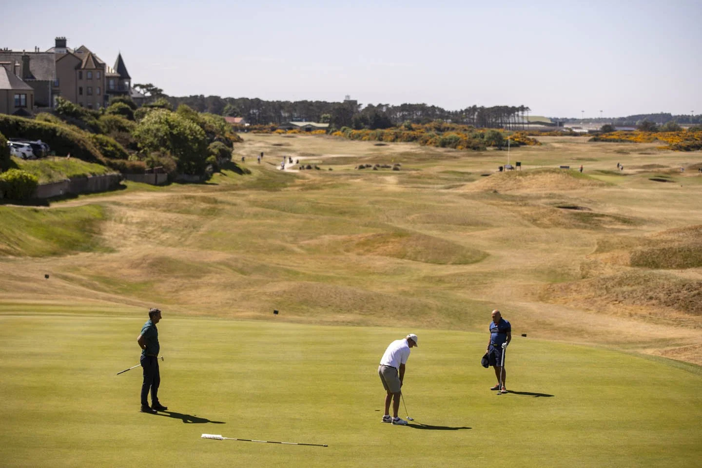 Three men playing golf on a green field with residential houses and rolling terrain in the background.