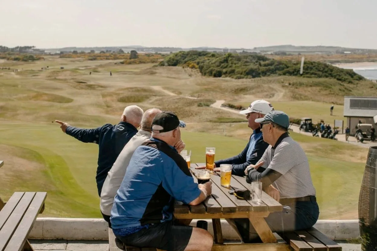 Group of men sitting at a picnic table outdoors, watching a golf course, with glasses of beer on the table.