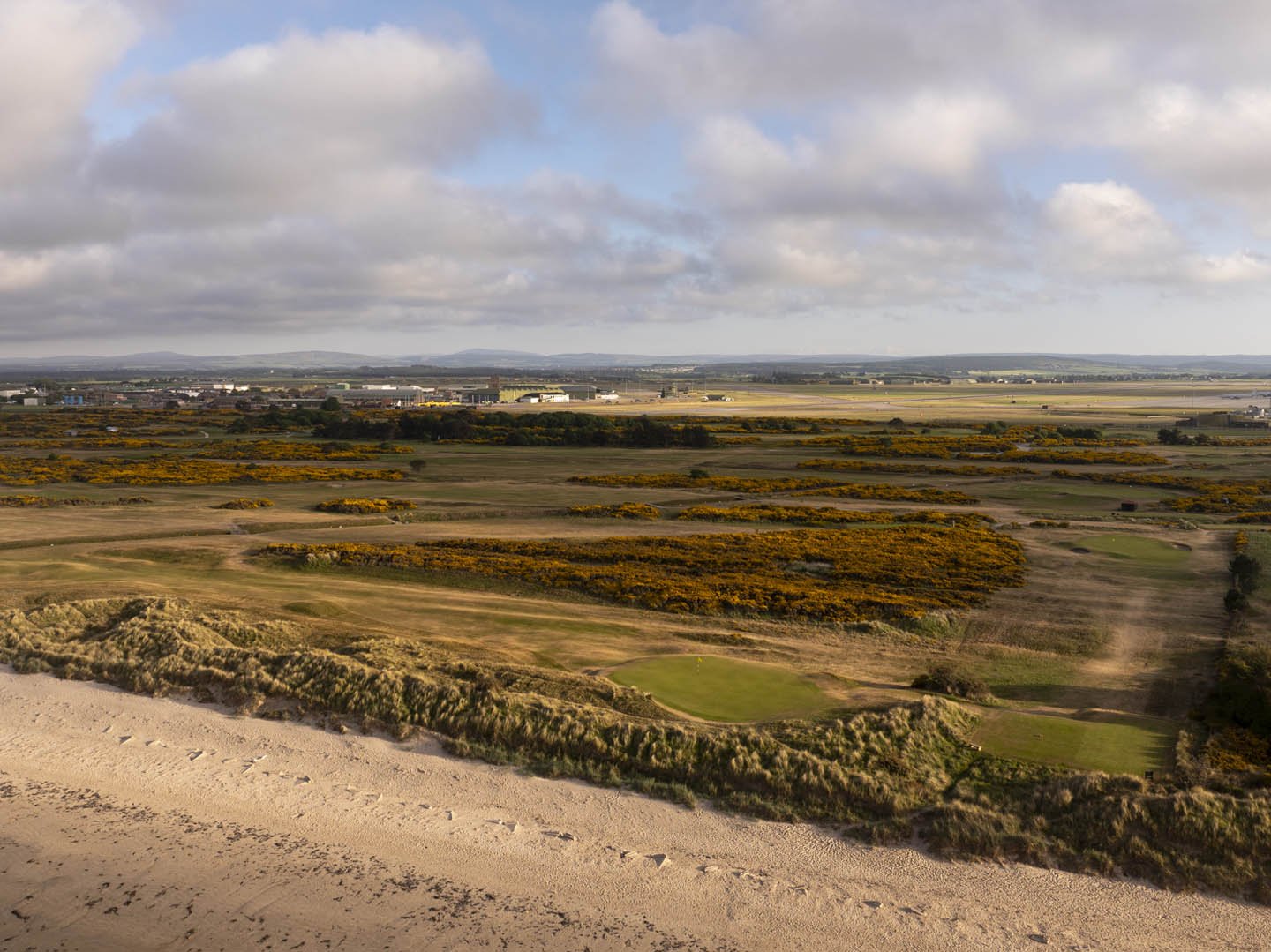 A landscape view of a golf course with sand bunkers and grassy areas, surrounded by yellow and green shrubbery, under a cloudy sky.