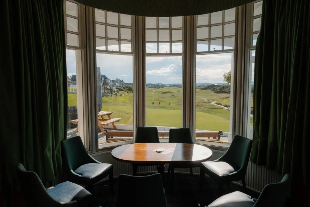 Room with a round table and six chairs, large bay window overlooking a green golf course and scenic landscape, with outdoor benches and picnic table visible outside.