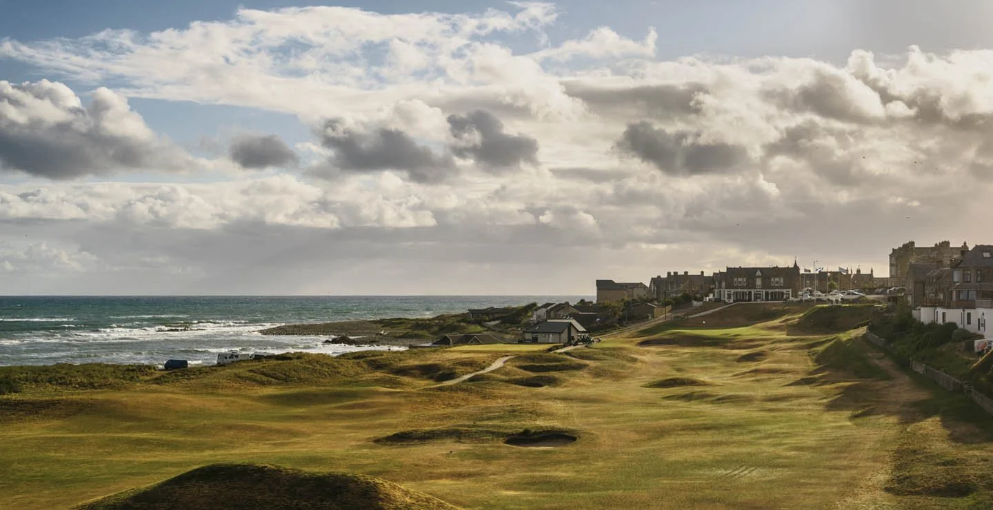A coastal golf course with sandy and grassy terrain near houses, overlooking the ocean under cloudy skies.