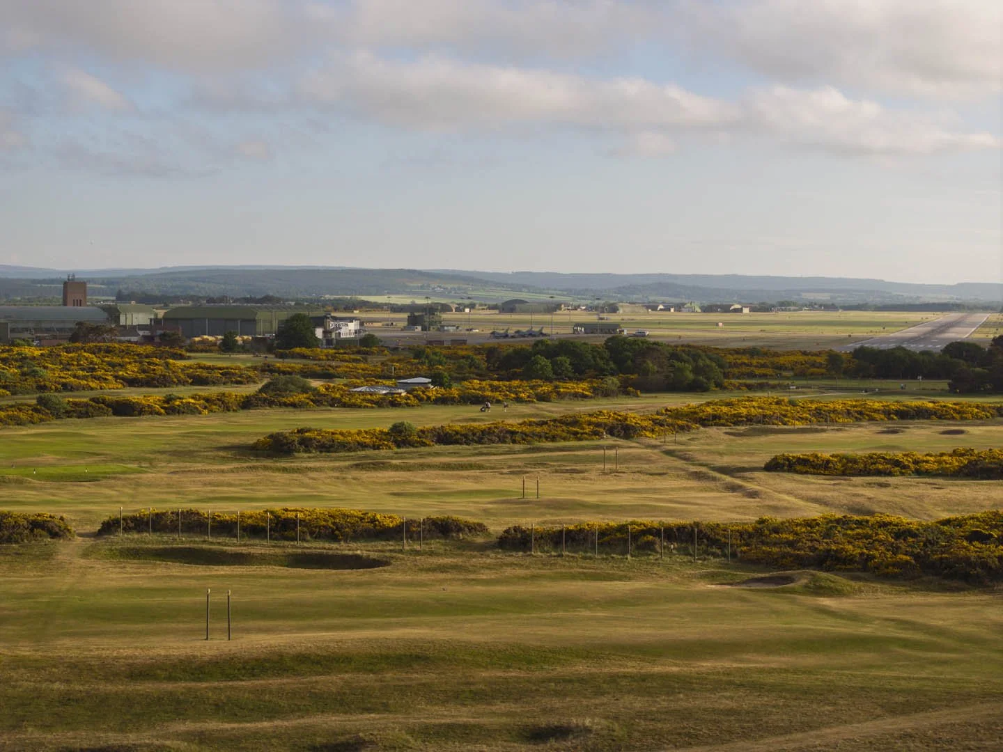 A wide view of a golf course with green grass, yellow flowering bushes, and paths, set against an industrial and rural landscape with buildings, roads, and distant hills under a partly cloudy sky.