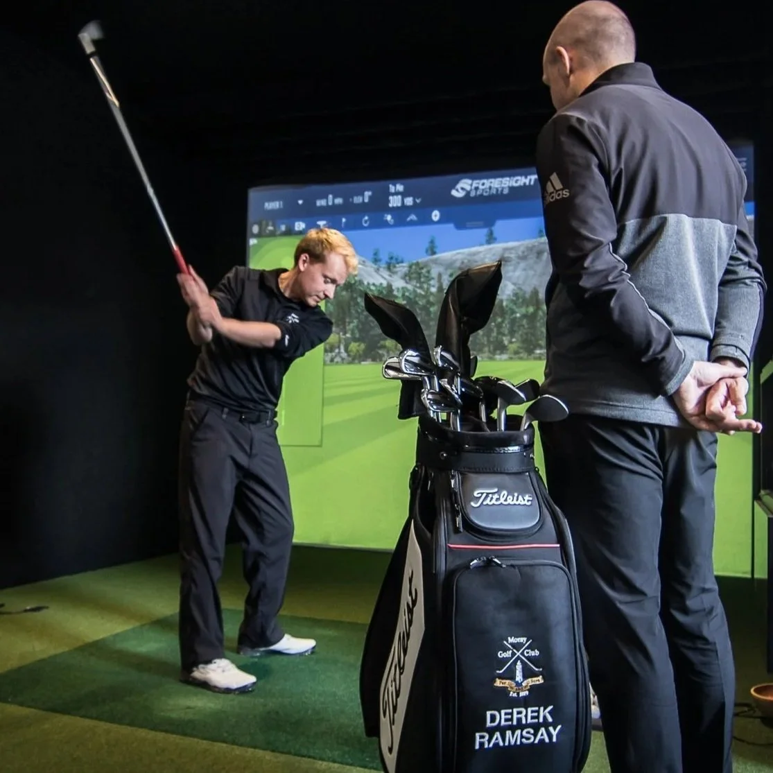 A man practicing golf swings in a simulator with a golf coach observing, inside a golf simulation room with a golf bag bearing the name Derek Ramsay.
