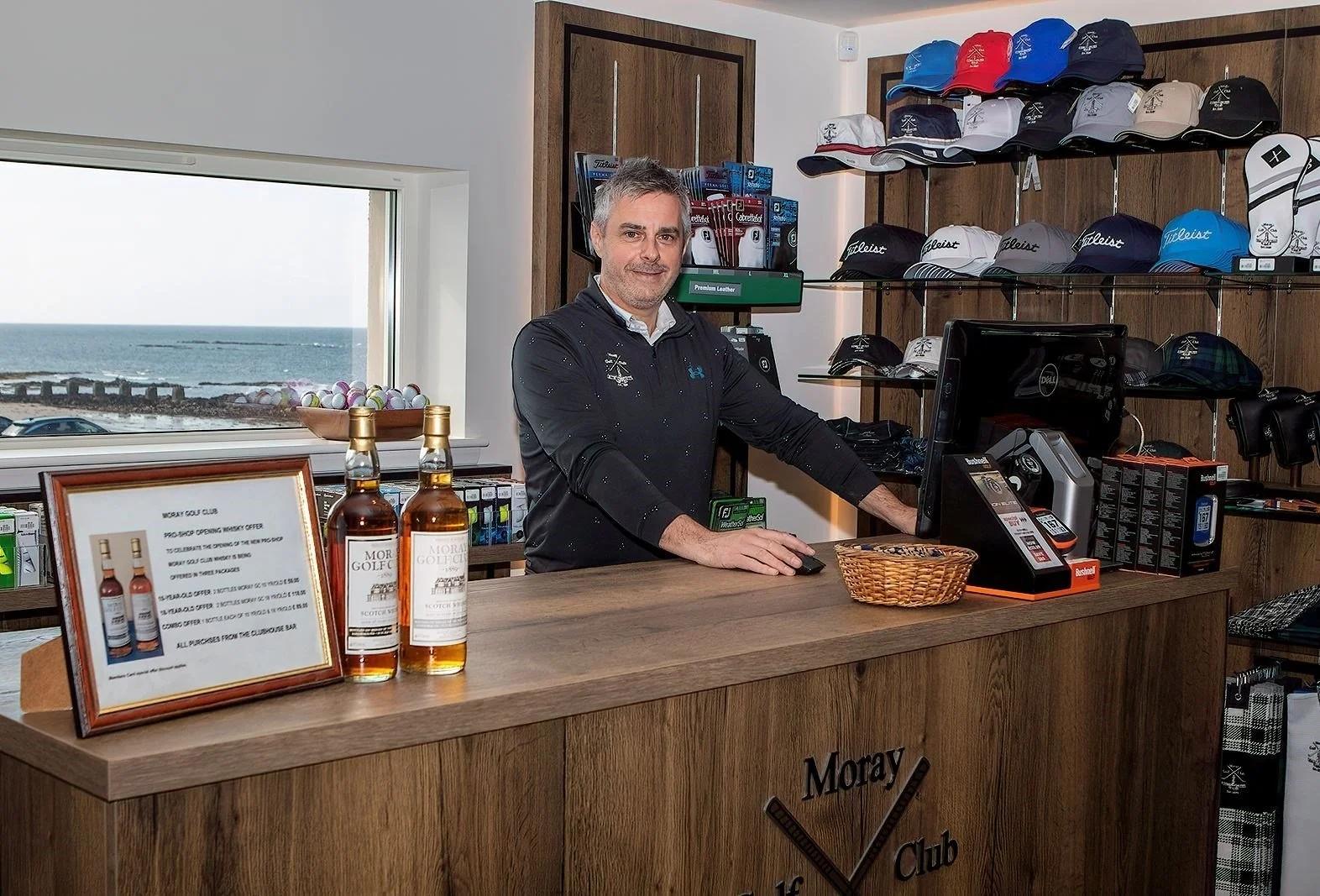 A man standing behind a wooden counter at a golf club, with golf caps on shelves behind him, a computer and a cash register in front of him, and a window showing the ocean in the background.