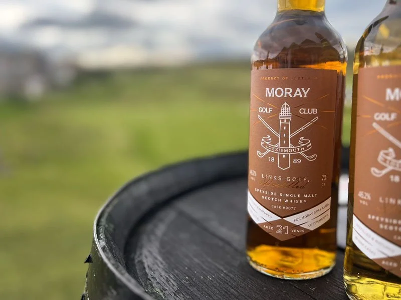 Close-up of a bottle of Moray single malt Scotch whisky with a golf theme label, placed on a black surface outdoors, with a blurred green landscape and cloudy sky in the background.