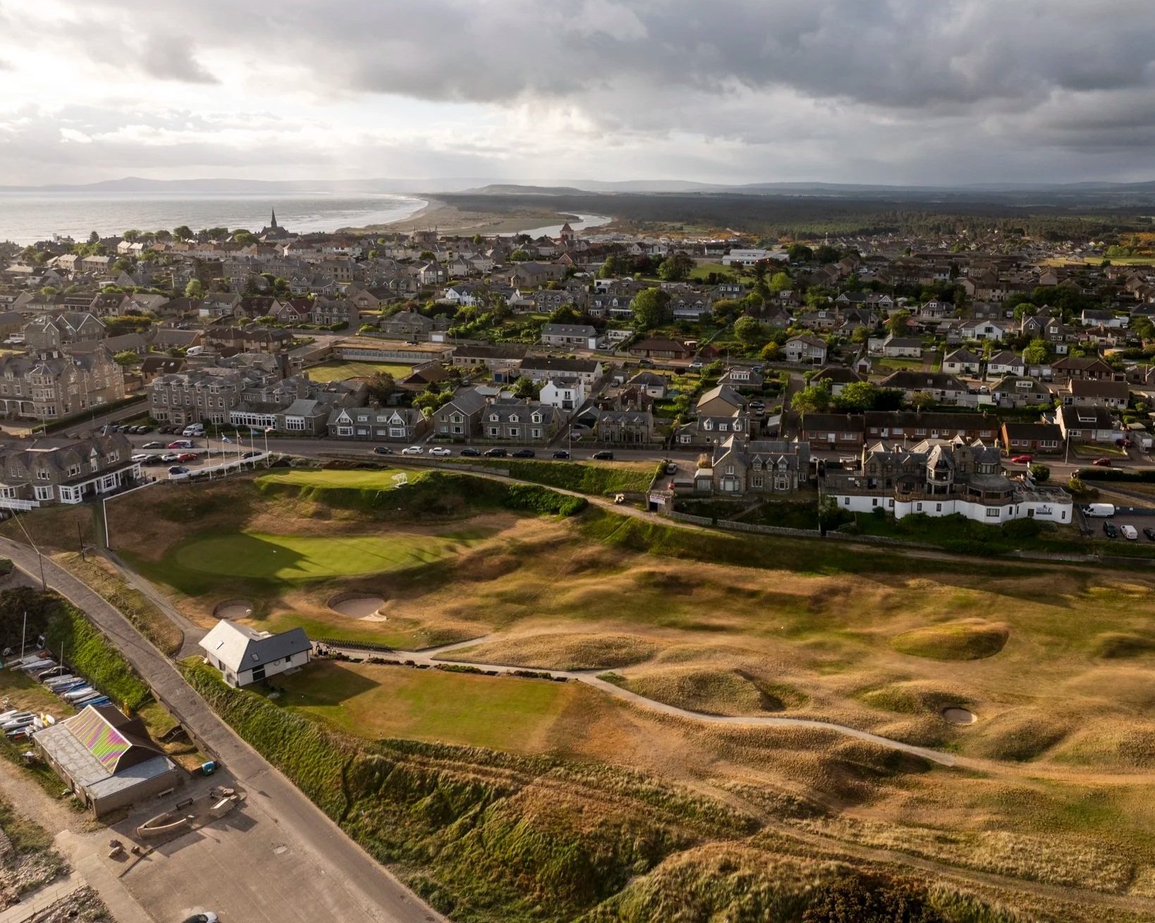 Aerial view of a coastal town with a golf course in the foreground and residential houses extending to the shoreline.