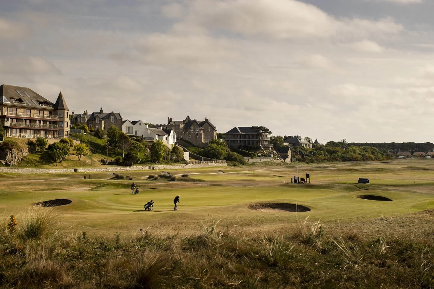 Golf course with three golfers playing near sand bunkers, residential houses and trees on a hillside in the background under a partly cloudy sky.