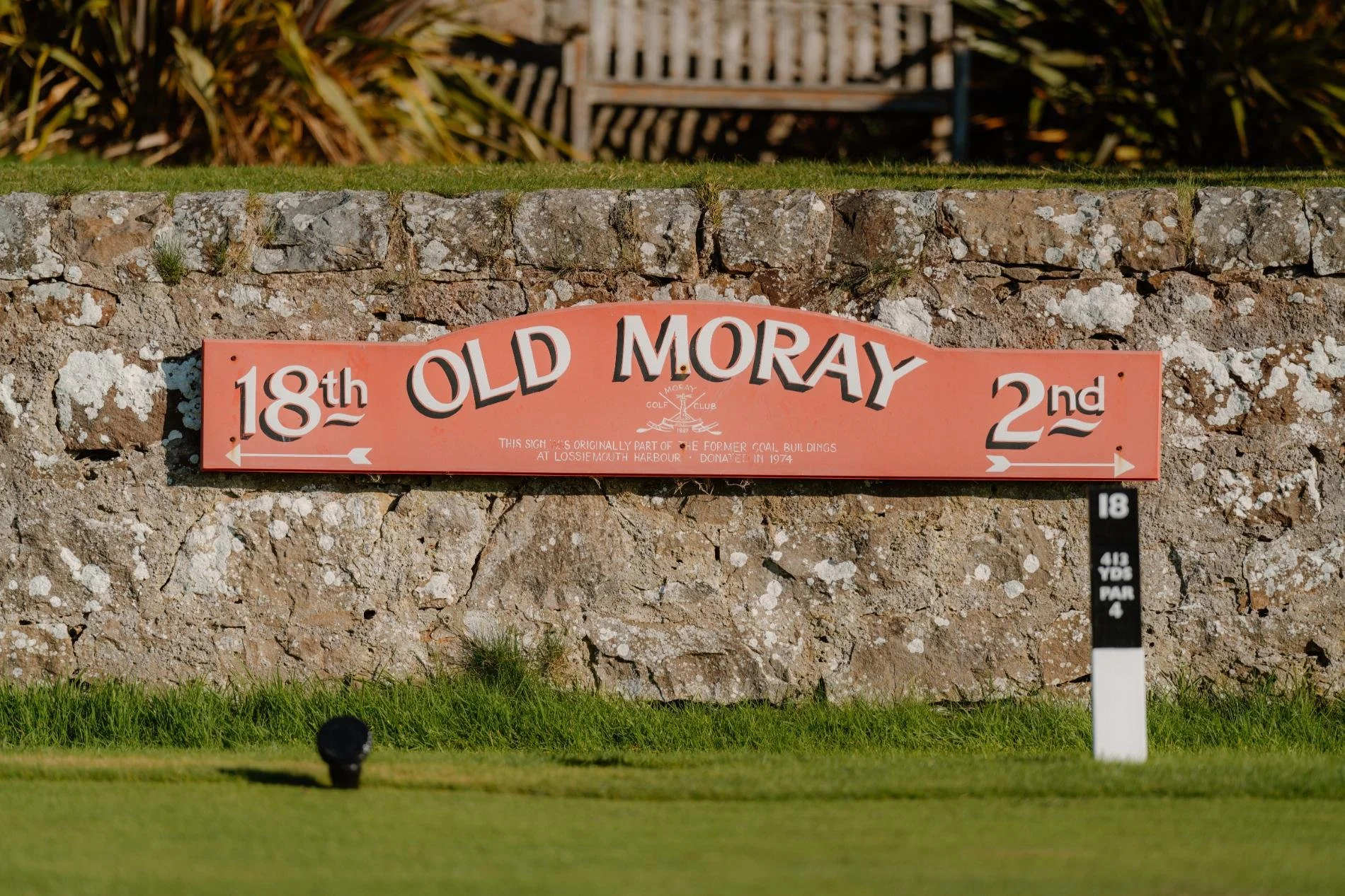 Golf course sign for Old Moray, indicating hole 18th to the left and 2nd to the right, with a stone wall and bench in the background.