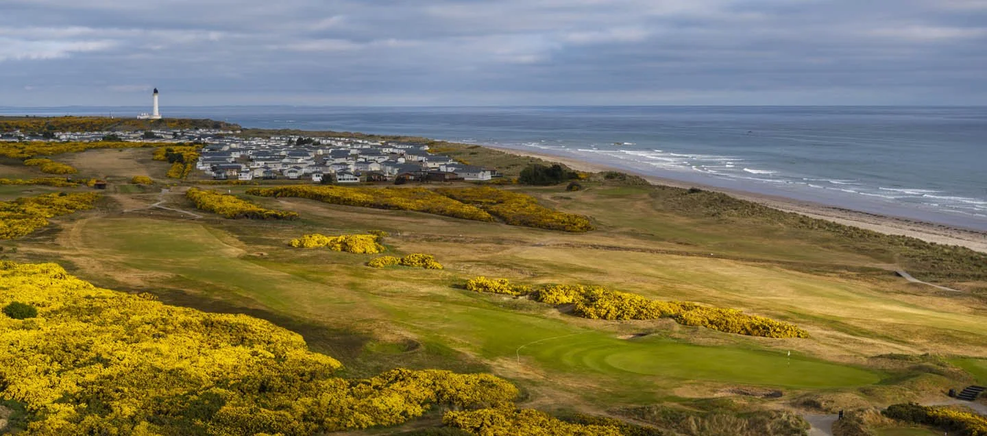 A scenic view of a golf course with yellow flowering bushes, a grassy fairway, a residential area, a lighthouse, and the ocean in the background under cloudy skies.