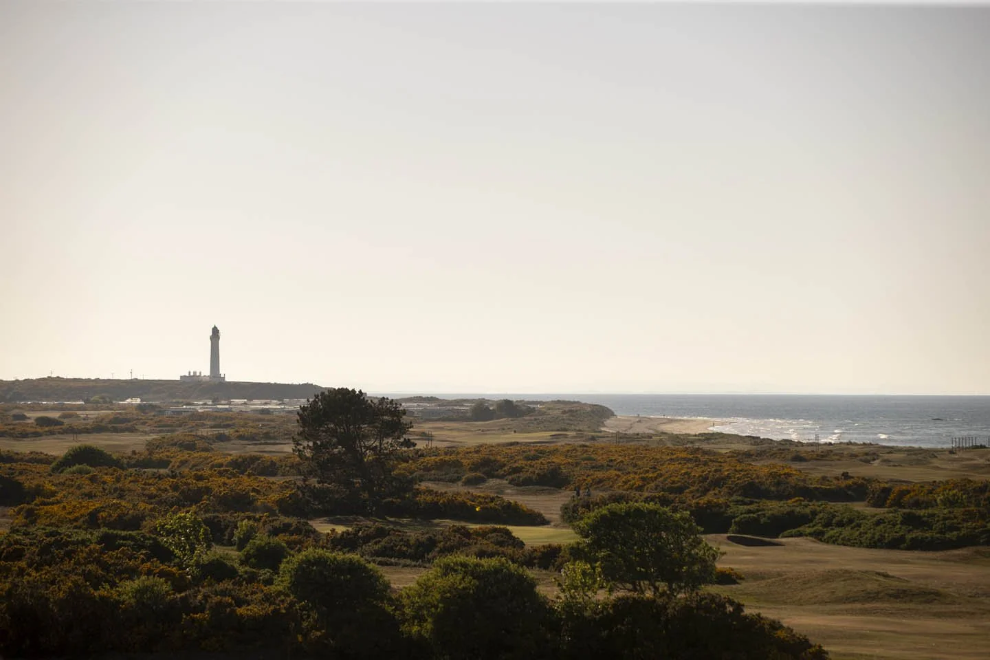 A coastal landscape with a golf course in the foreground, trees along the fairways, and a lighthouse on a distant hill near the shoreline under a clear sky.