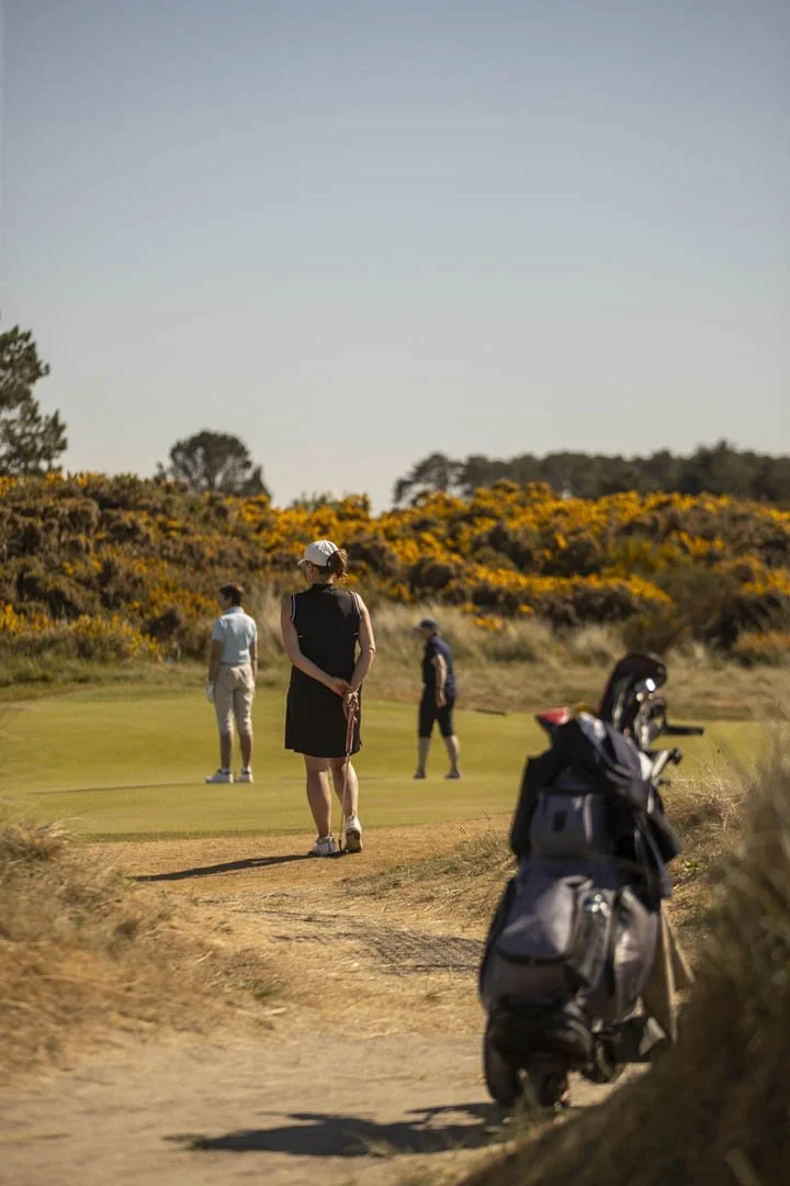 People playing golf on a course with yellow flowering bushes in the background and a golf bag in the foreground.