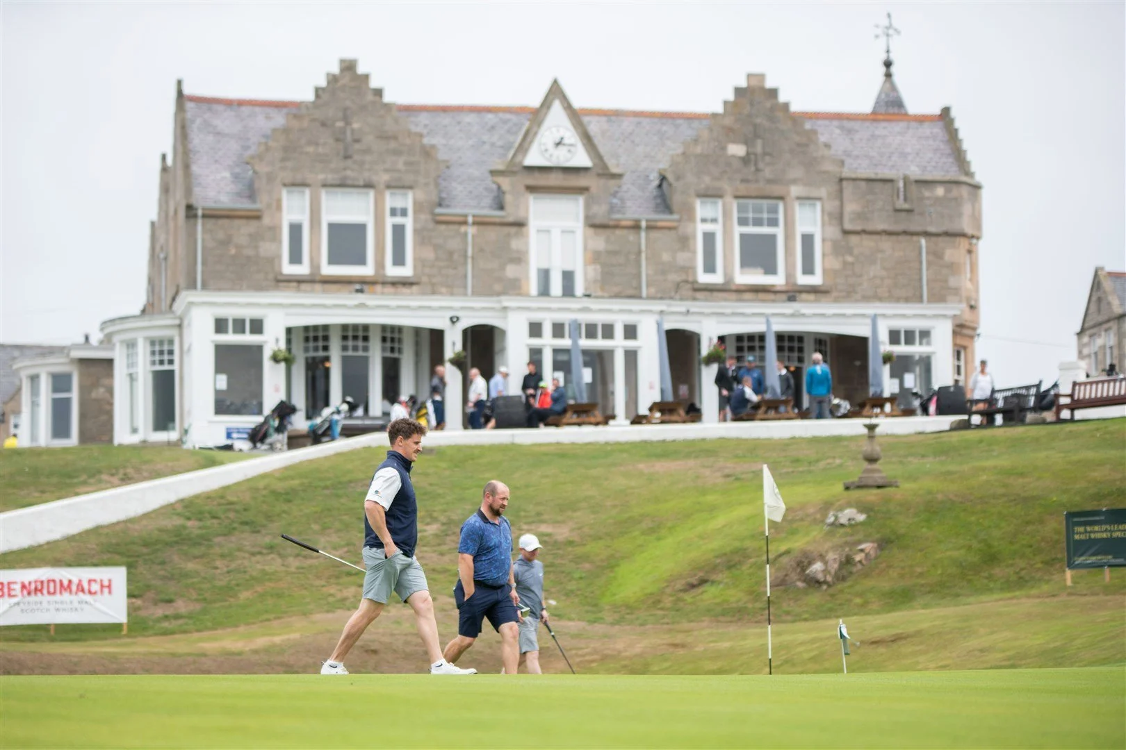 People playing golf on a green, with a large historic building and a restaurant patio in the background.