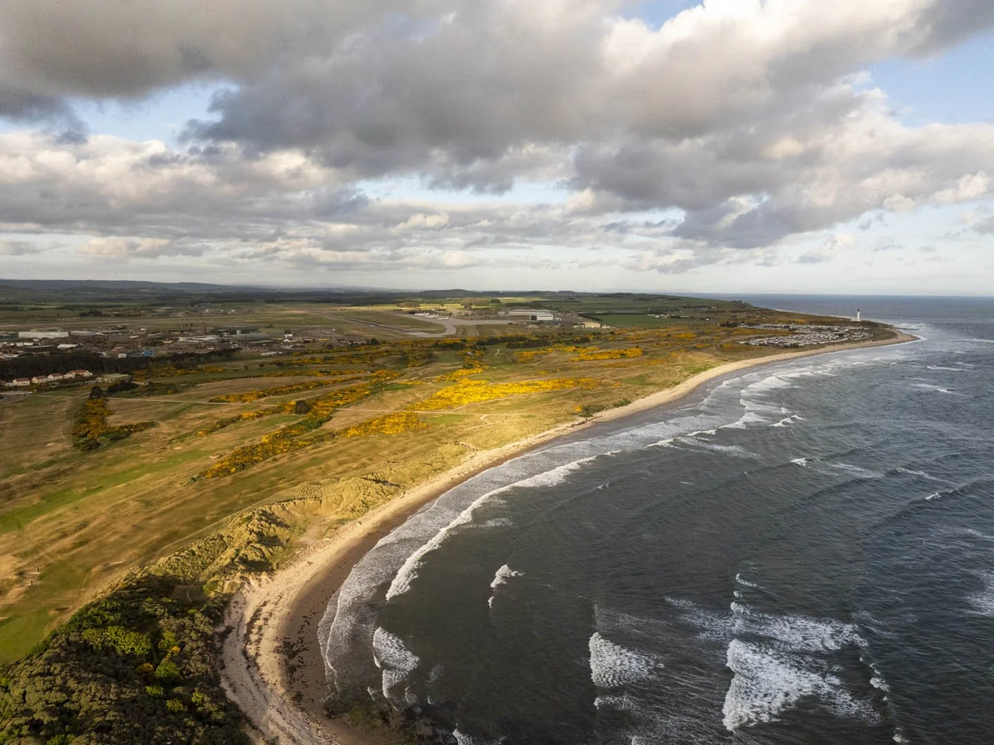 Aerial view of a coastline with a sandy beach, grassy cliffs, and yellow-flowered bushes, with ocean waves crashing onto the shore under a partly cloudy sky.