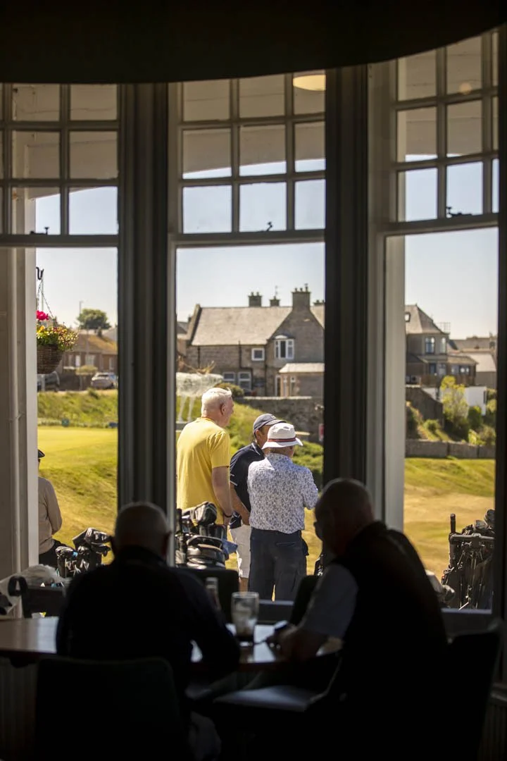 People inside a windowed room watching golfers on a golf course outside.