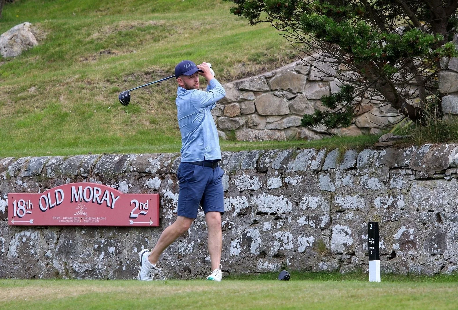 A man in a blue shirt, navy shorts, and a navy cap preparing to hit a golf ball on the sidewalk near a stone wall, with a golf course sign for the 18th hole.