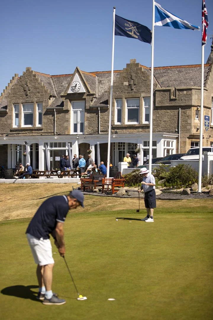 People playing golf on the putting green at a golf club, with a stone clubhouse and flags in the background on a sunny day.