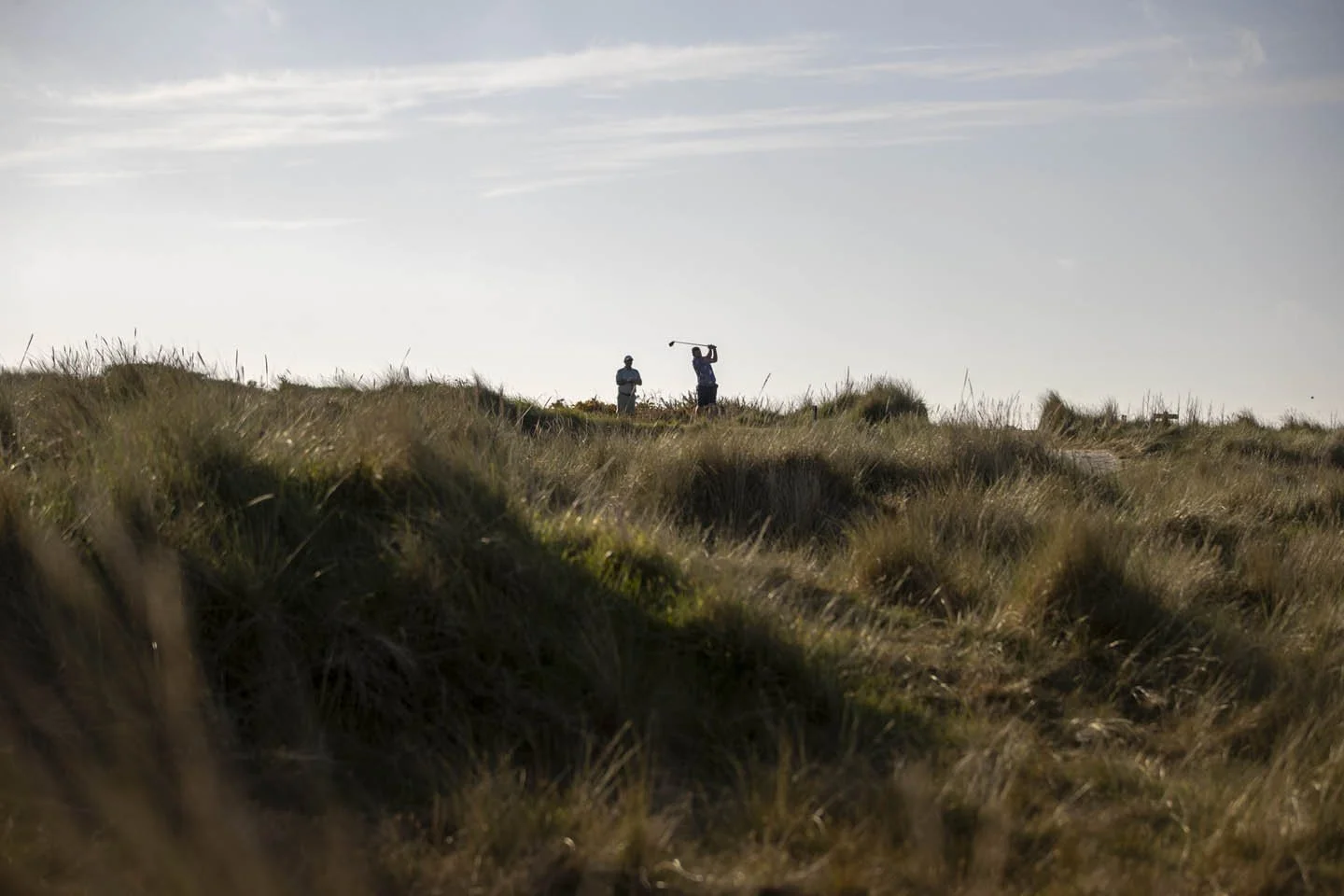 Two people standing in a grassy field, one holding a golf club in a backswing position, under a partly cloudy sky.