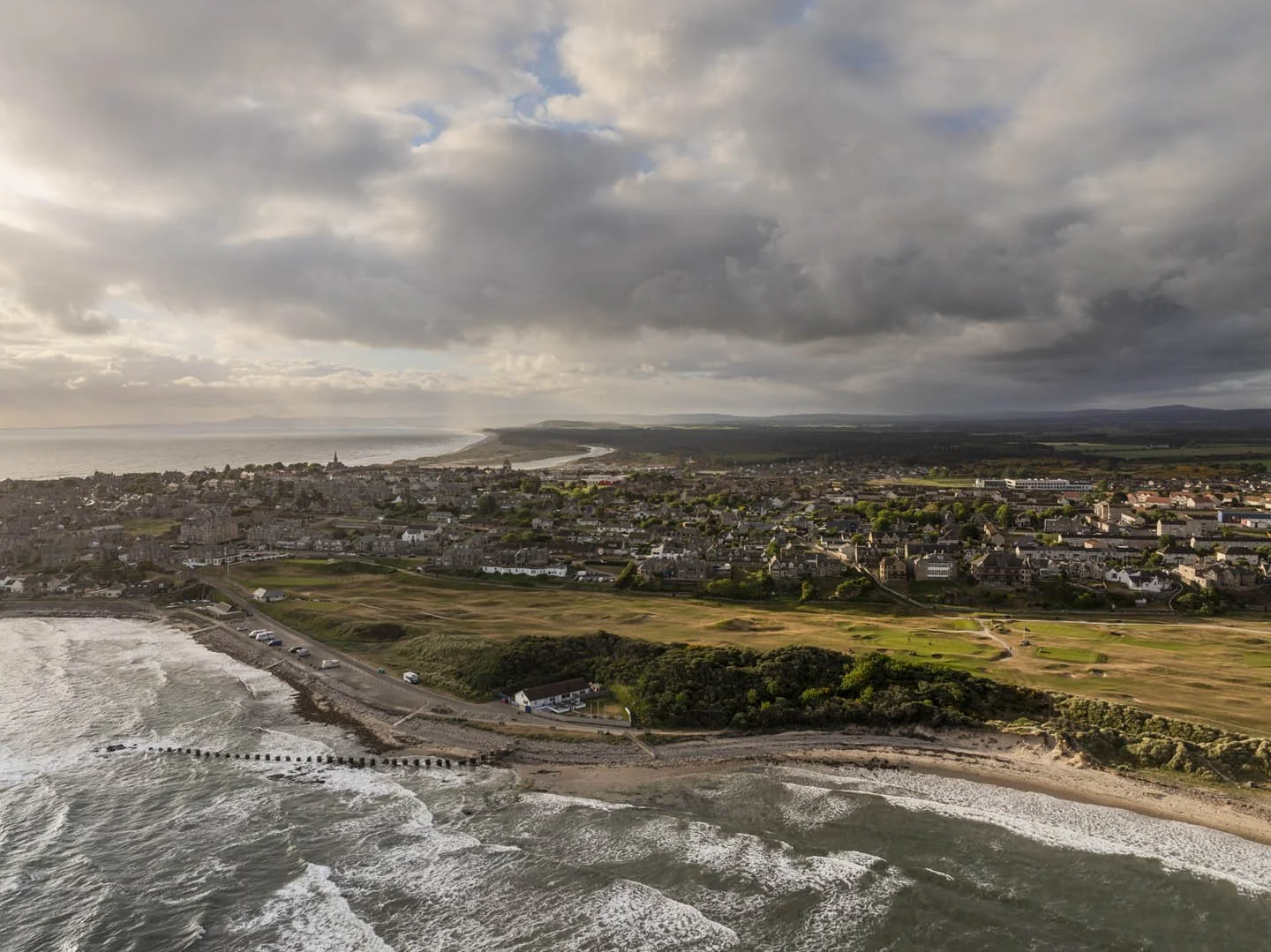 A coastal town with a beach in the foreground, a golf course, and residential buildings extending to the horizon under cloudy skies.