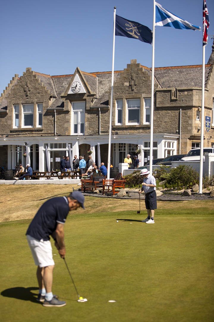 Two men playing golf on the green at a golf course, with a large stone building and flags in the background.
