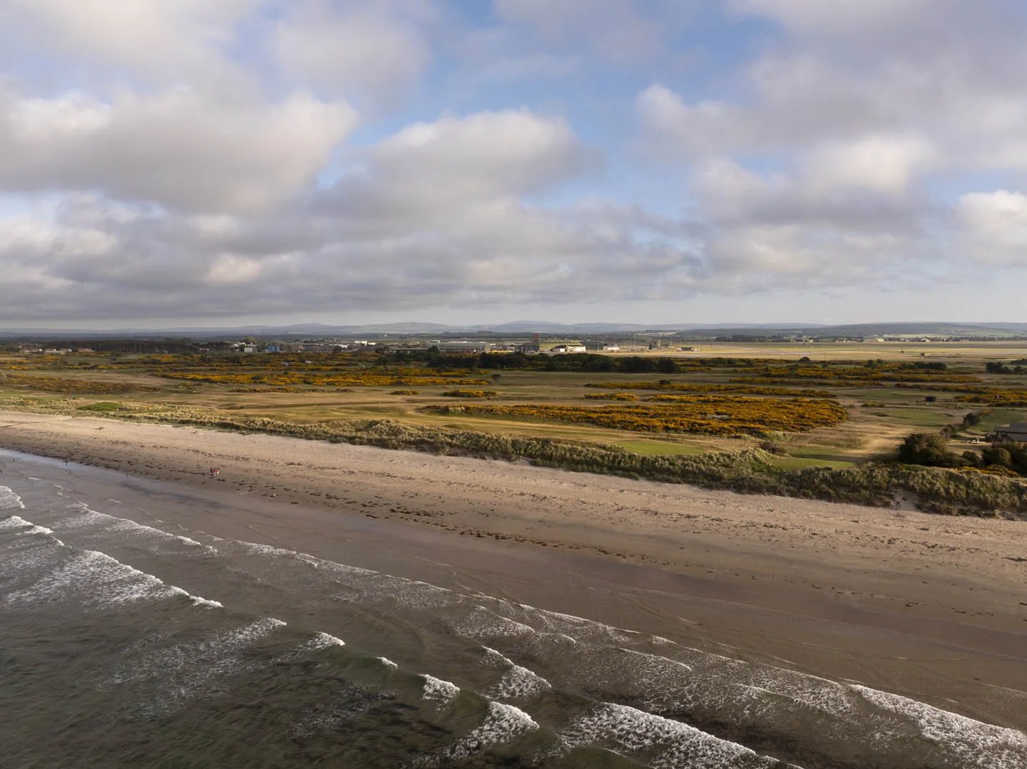 A view of a sandy beach with ocean waves, and a grassy, partly yellow-flowered landscape with some distant buildings and a cloudy sky overhead.