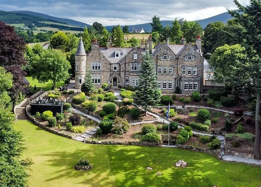 A large historic mansion with turret and multiple windows, surrounded by a landscaped garden with shrubs, trees, and stone pathways, situated in a lush green landscape with hills in the background.
