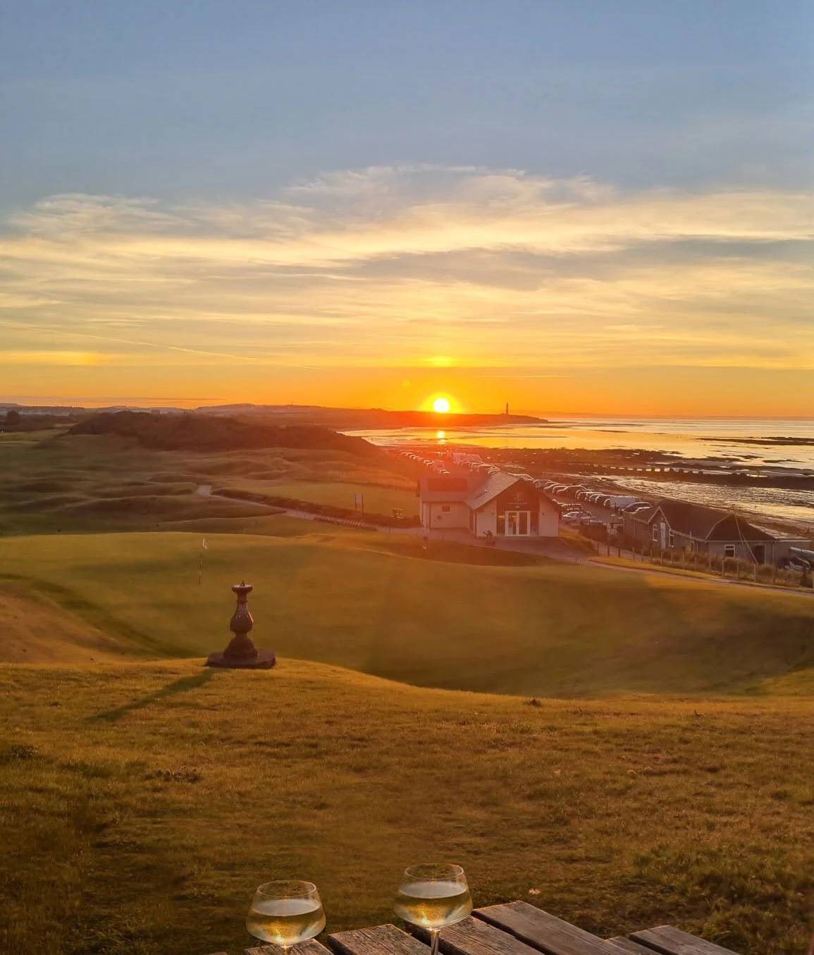 Sunset over a coastal golf course with a view of the ocean, houses along the shoreline, and two glasses of white wine on a wooden table in the foreground.