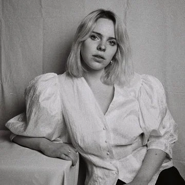 Black and white photo of a young woman with shoulder-length blonde hair, wearing a light-colored blouse with puffed sleeves, sitting against a plain background.