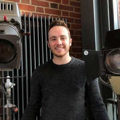 A young man with short brown hair and a beard, smiling, wearing a dark sweater, standing indoors near a large camera and studio equipment with a brick wall and radiator in the background.
