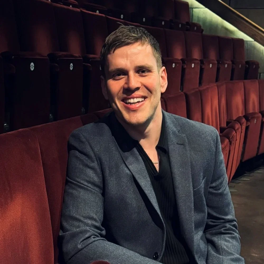 A young man smiling and sitting in an empty theater with red seats.