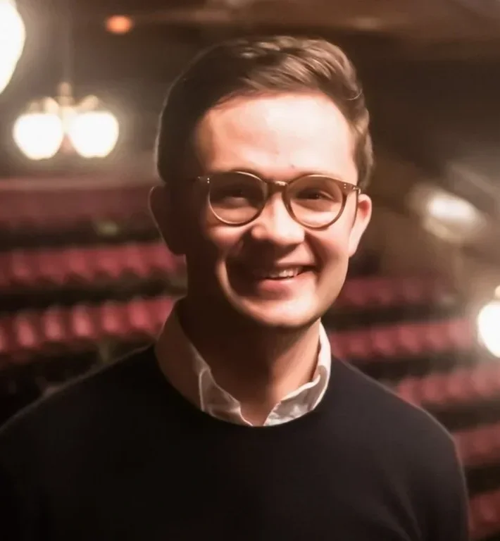 A young man with glasses smiling in a dimly lit theater or auditorium with red seats in the background.