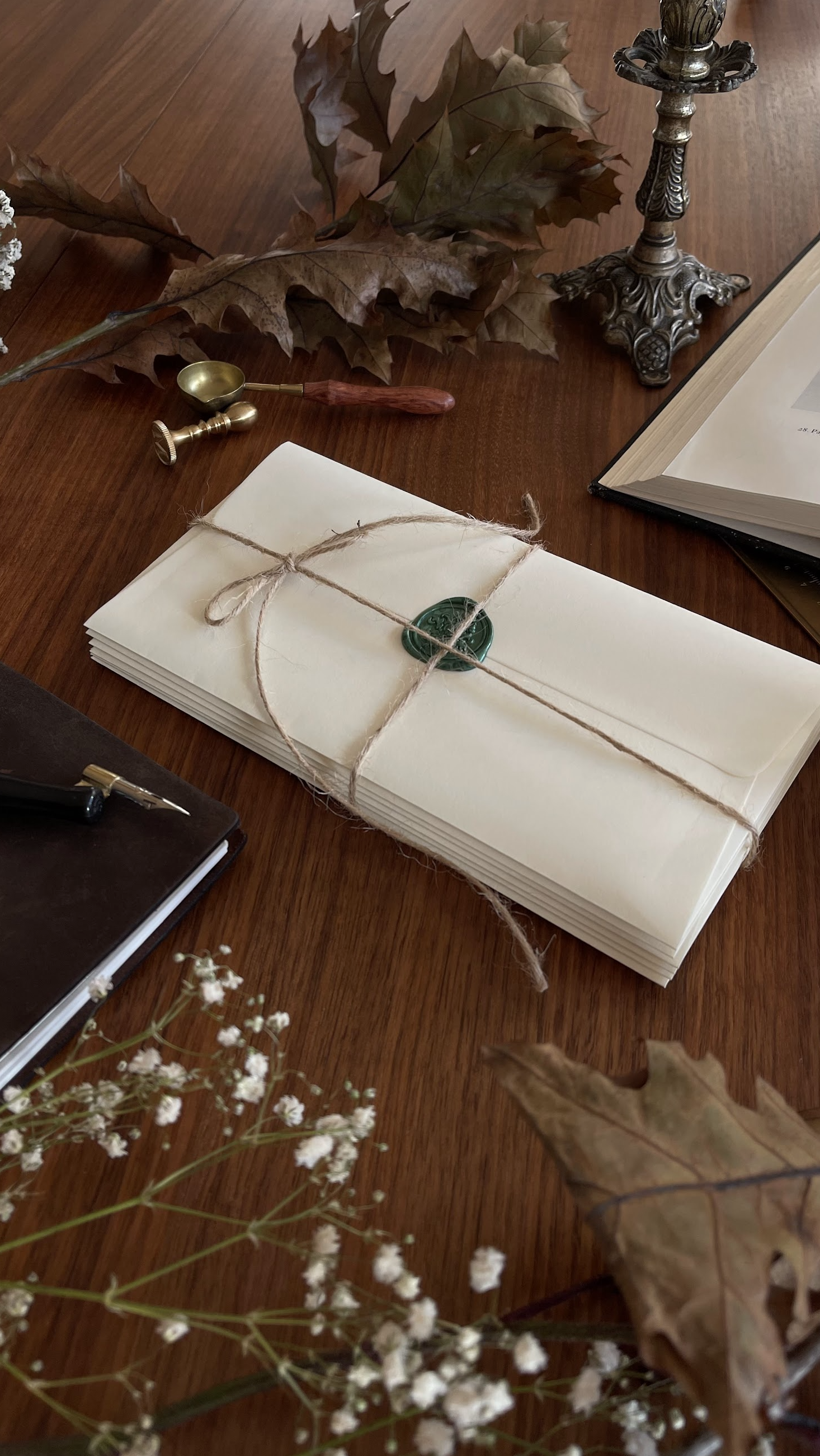 A bundle of cream-colored envelopes sealed with a green wax stamp, tied with twine, on a wooden table surrounded by dry autumn leaves, a small brass scoop and pestle, a lit candlestick, a black notebook, and white flowers.