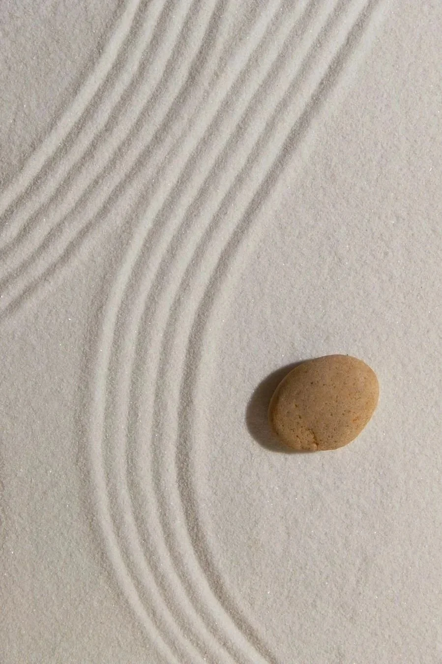 A small, light-brown stone on a smooth white surface with evenly spaced, curved lines drawn in the sand.