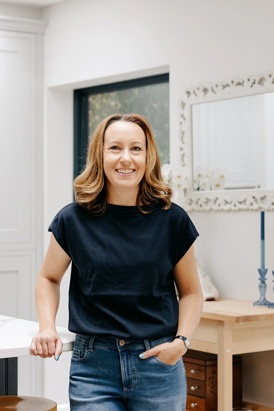 A woman with shoulder-length wavy brown hair, wearing a black t-shirt and jeans, smiling and standing in a well-lit room.