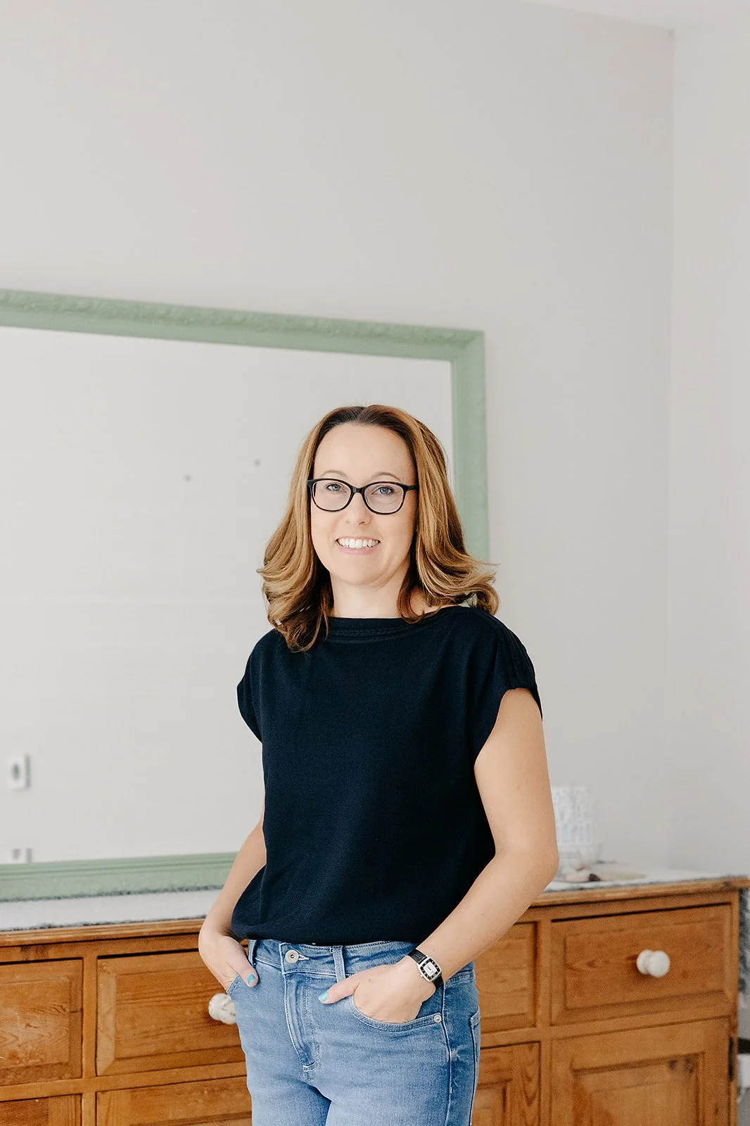 A woman with shoulder-length light brown hair, wearing glasses, a black T-shirt, and blue jeans, standing indoors in front of a mirror with green trim and a wooden cabinet.