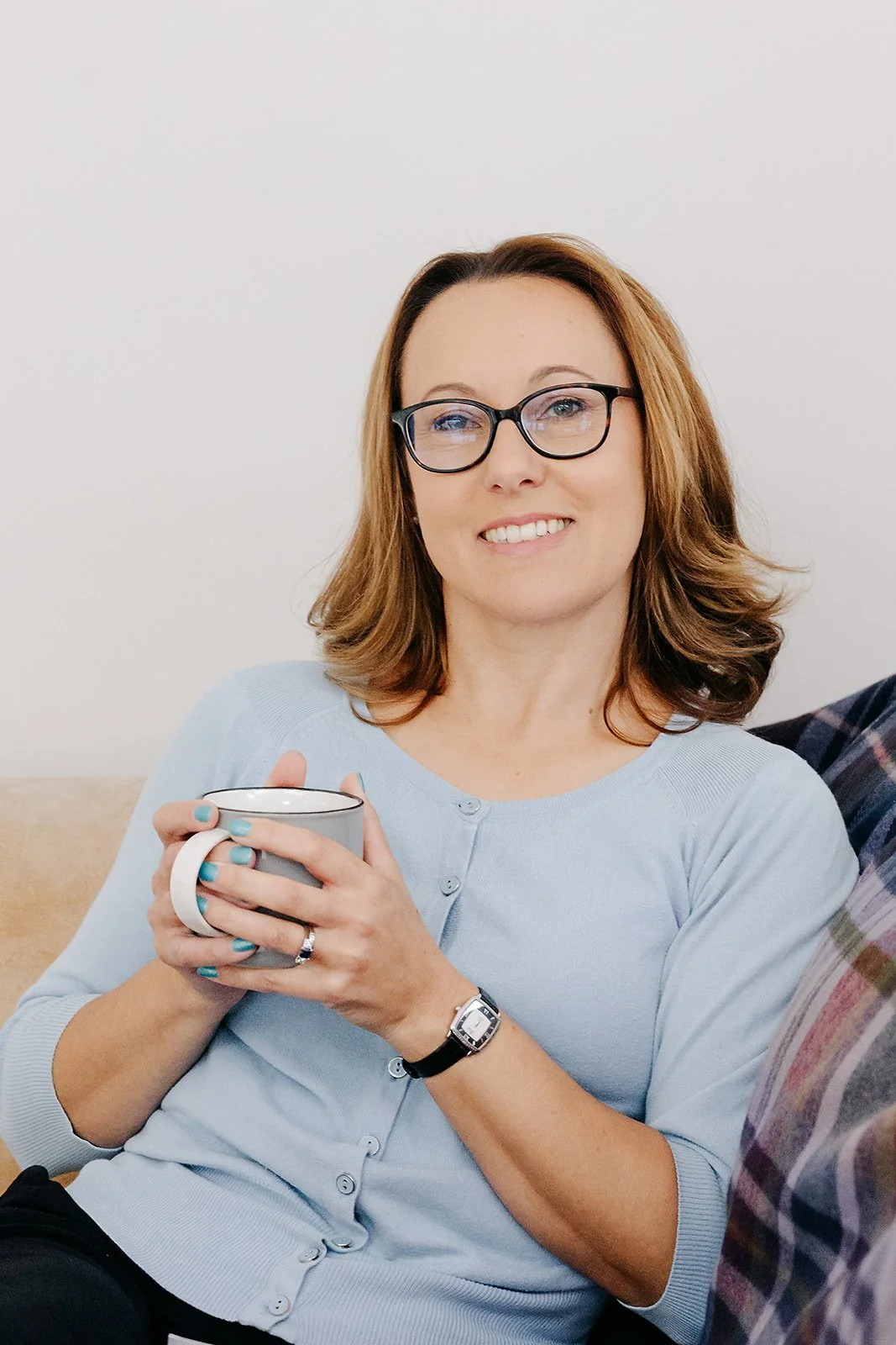 Woman with glasses holding a coffee mug, sitting on a beige couch, smiling at the camera.