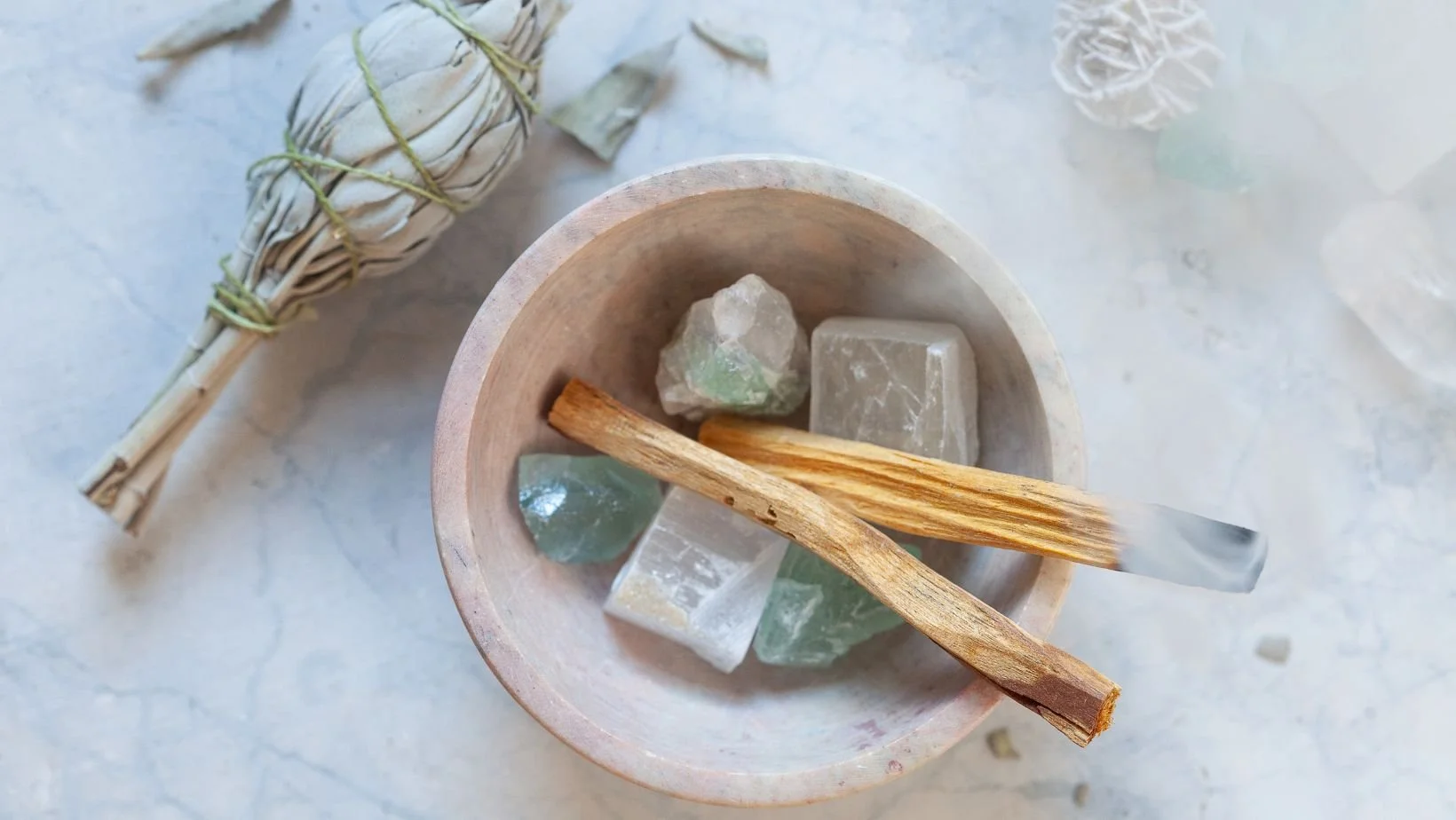A wooden bowl holds six pieces of various stones and crystals, including some with pale green and clear coloration, with two wooden sticks resting across the stones. A bundle of dried white herbs or flowers tied with string is positioned beside the bowl on a light surface.