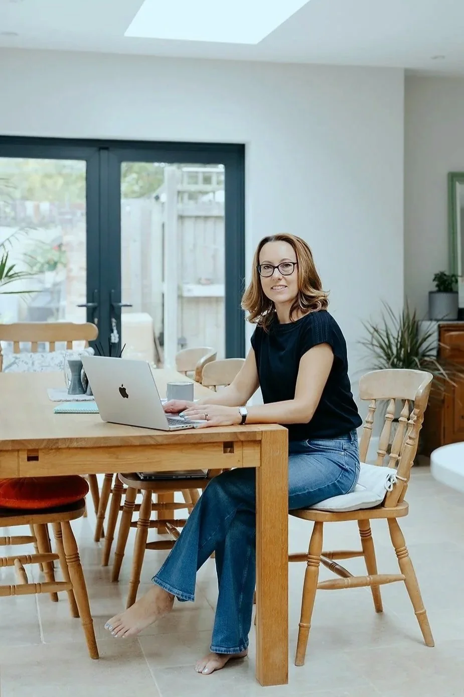 A woman with glasses, sitting barefoot at a wooden table in a bright, modern dining area with a laptop, mug, and notebook, near large glass doors leading to an outdoor space.