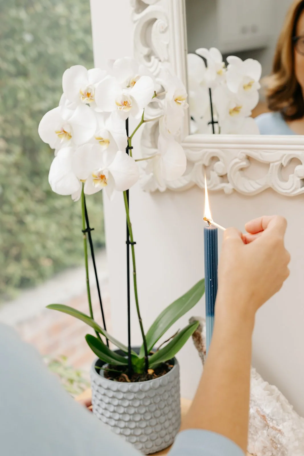 A person is lighting a blue candle with a matchstick near a white orchid plant in a textured pot, against a mirror with ornate white frame.