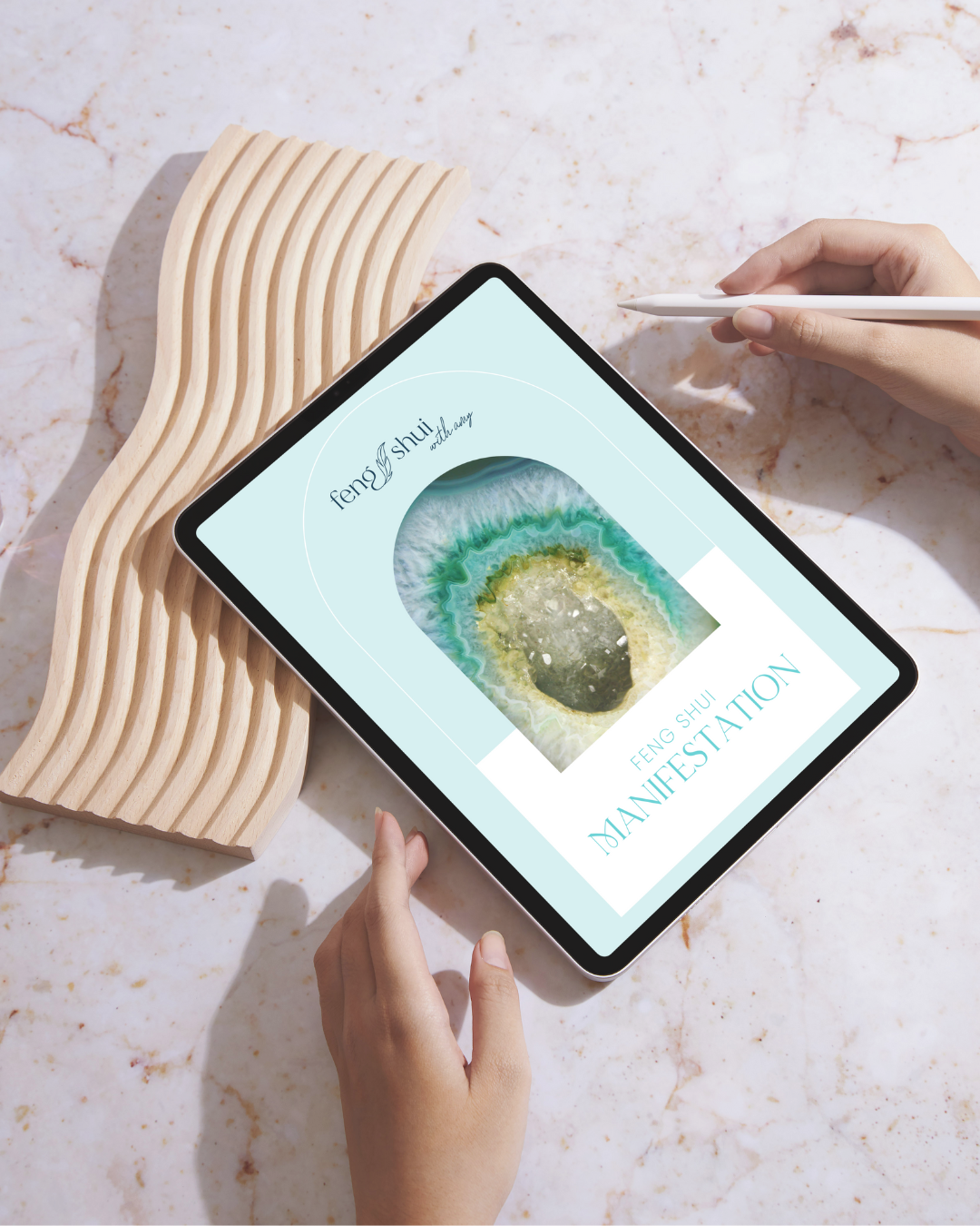 A person holding a tablet with a digital manifestation of a Feng Shui design featuring a circular stone or mineral. The tablet is resting on a white marble surface with a wavy wooden decorative piece beside it.