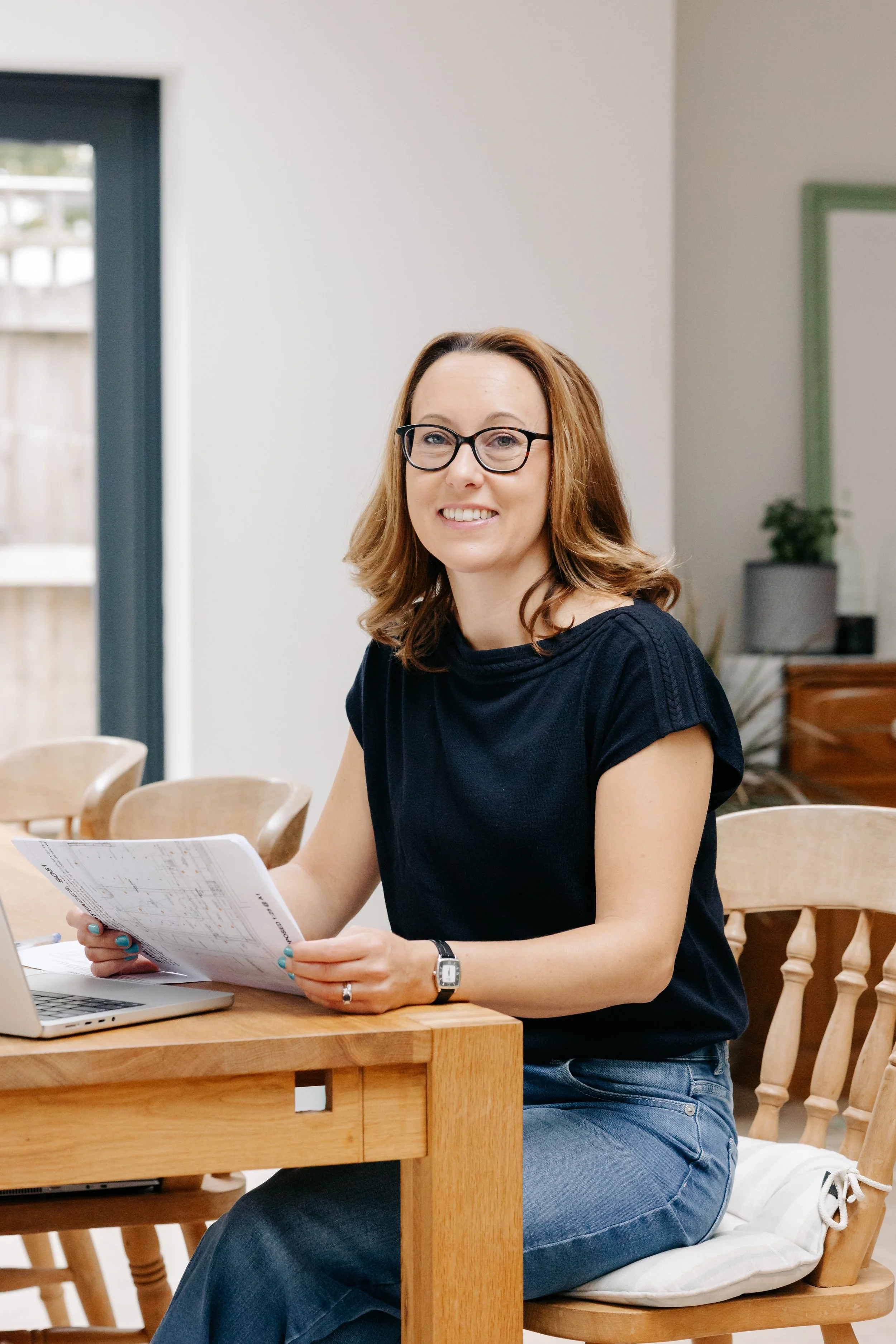 A woman with shoulder-length red hair, glasses, and a navy blue T-shirt sitting at a wooden table in a bright room, holding some papers and smiling at the camera.